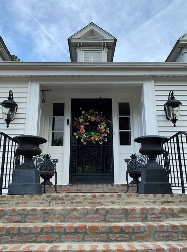 A white house entrance with brick stairs, black planters, ornate black metal railings, and a colorful floral door wreath.