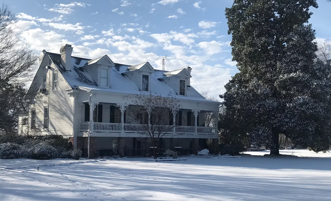 A white two-story historic house with a wraparound porch and dormer windows, surrounded by snow and a large evergreen tree.