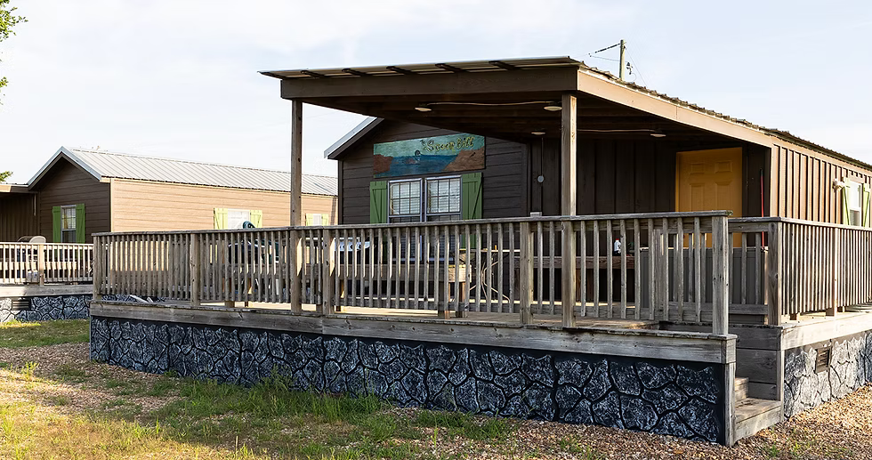 Two brown modular cabins with large wooden decks and patterned stone skirting sit in a grassy area under a bright sky.