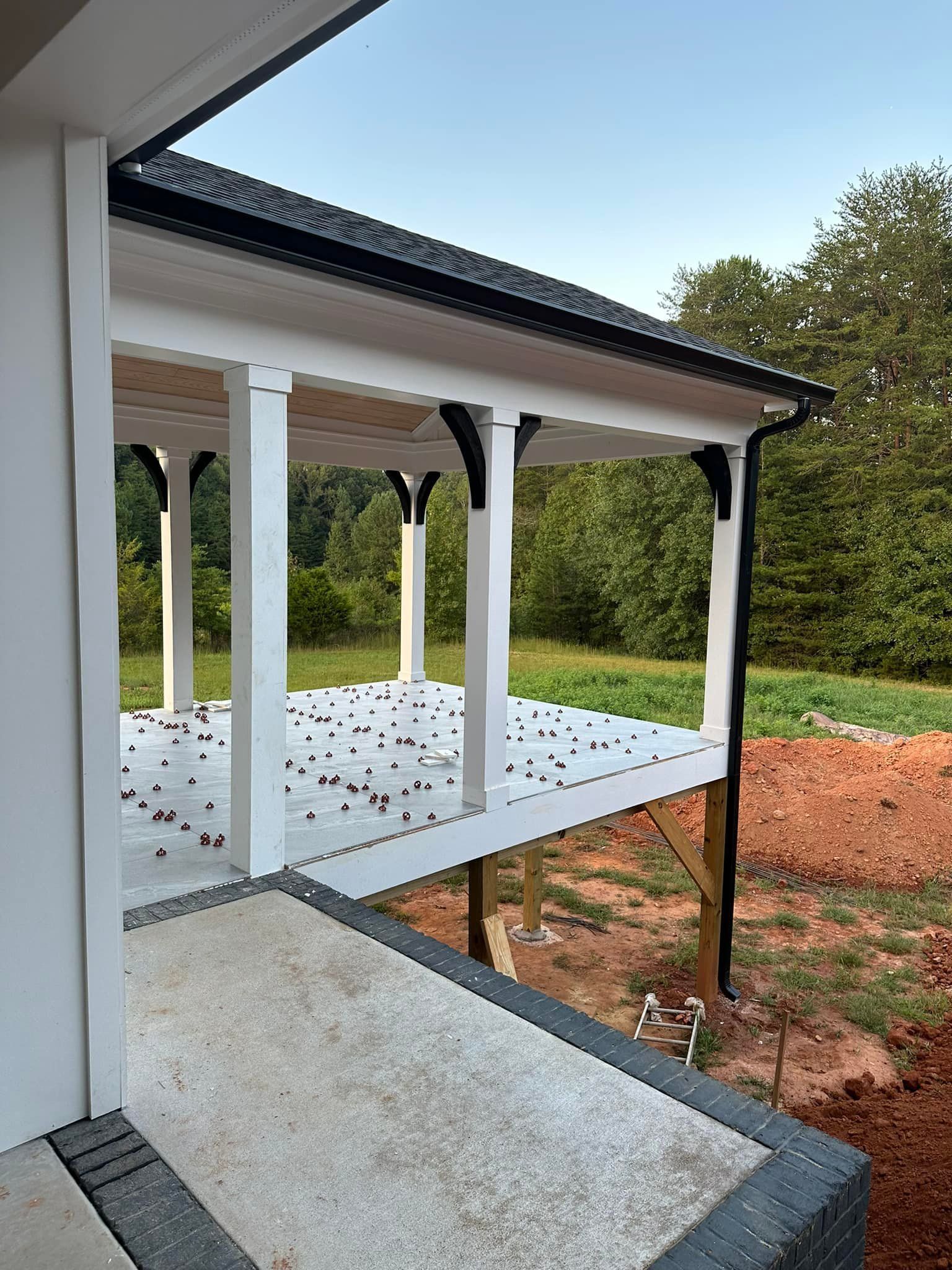 Covered porch with white pillars, black trim, and a gray concrete floor, overlooking a wooded area.