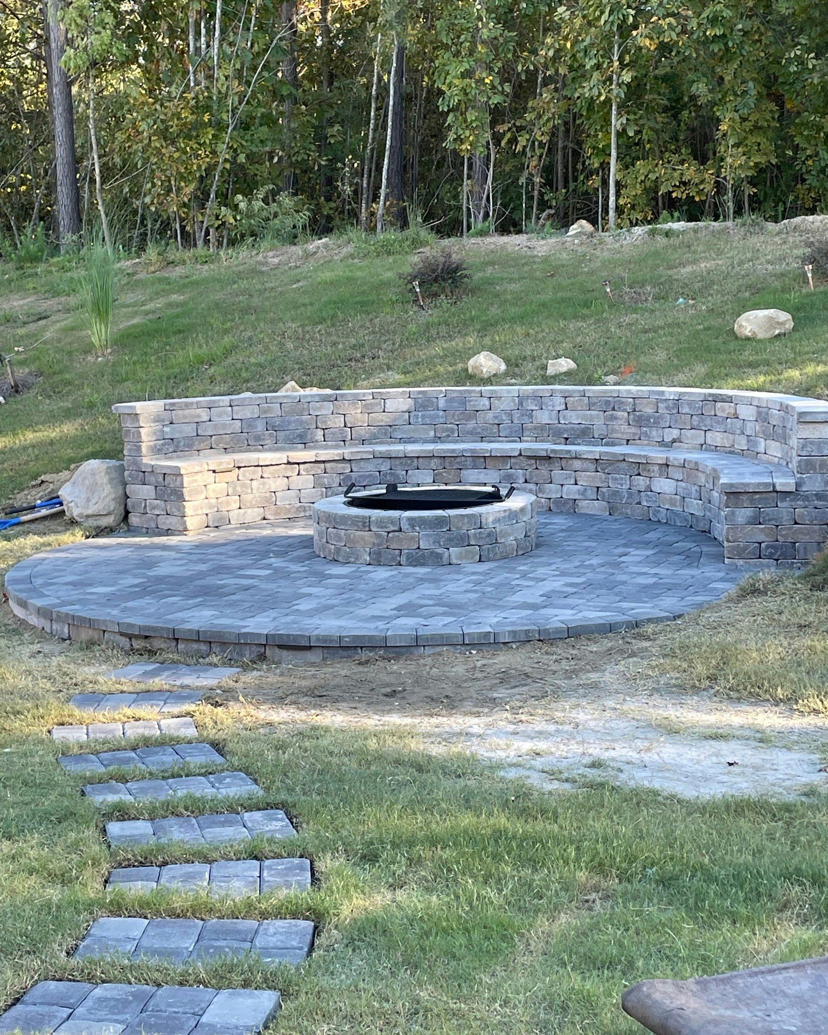 Stone patio with built-in seating around a fire pit on a grassy hillside, with a stone path leading to it.
