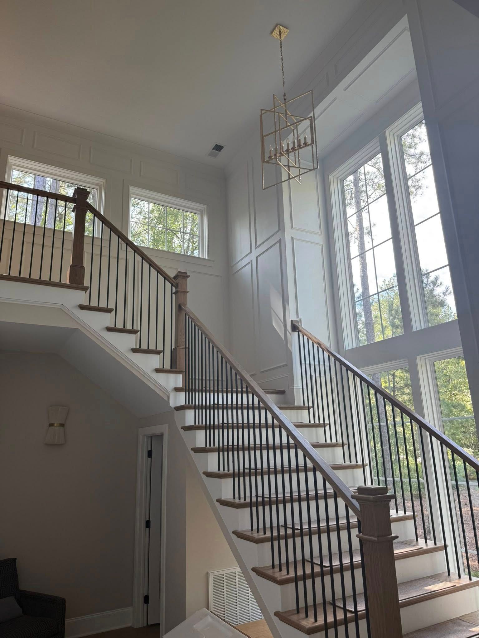 Staircase with white risers, dark spindles, and a chandelier; tall windows in the background.