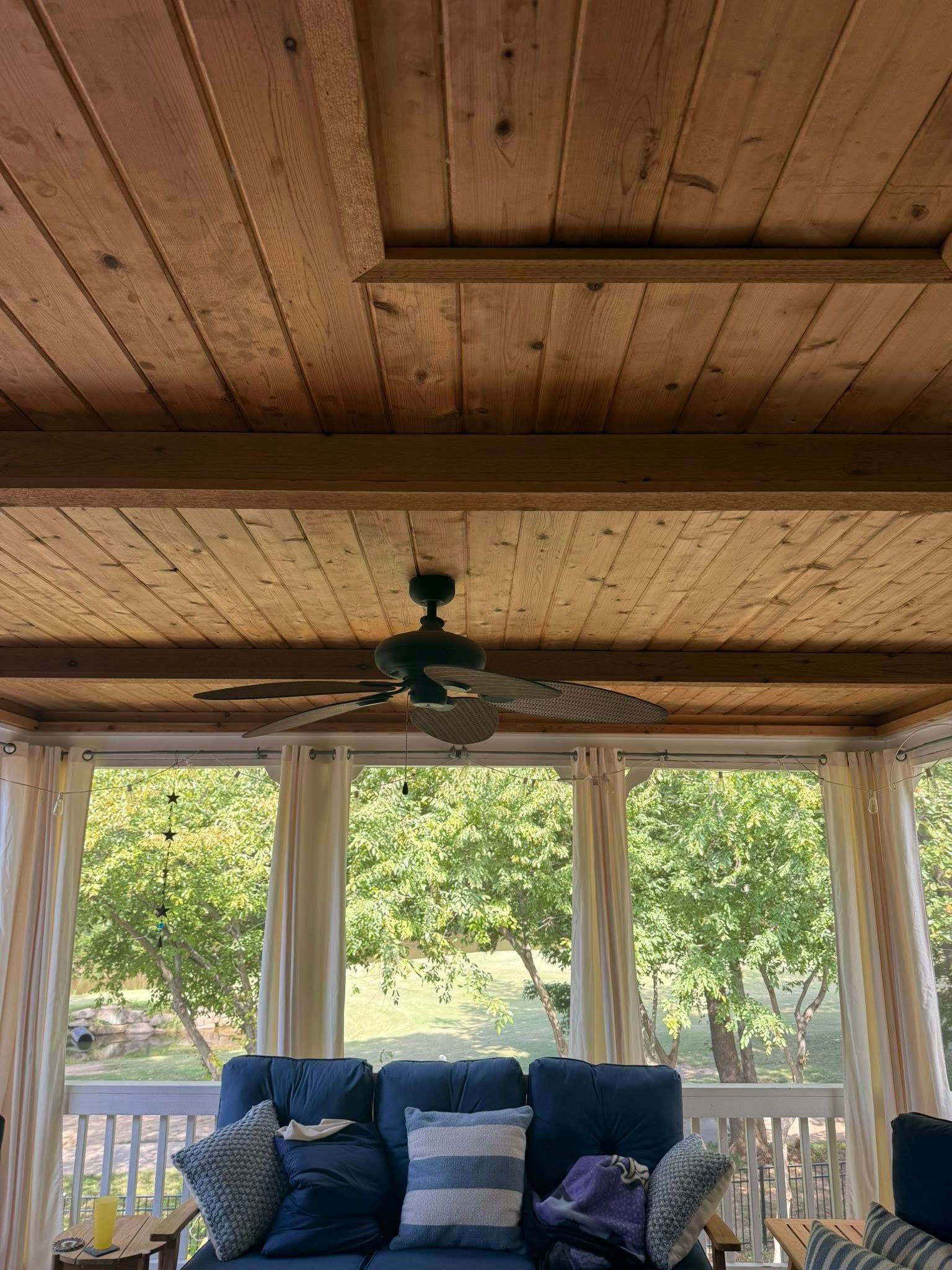 Screened porch with wood ceiling and beams, ceiling fan, blue couch, white curtains, and outdoor view.