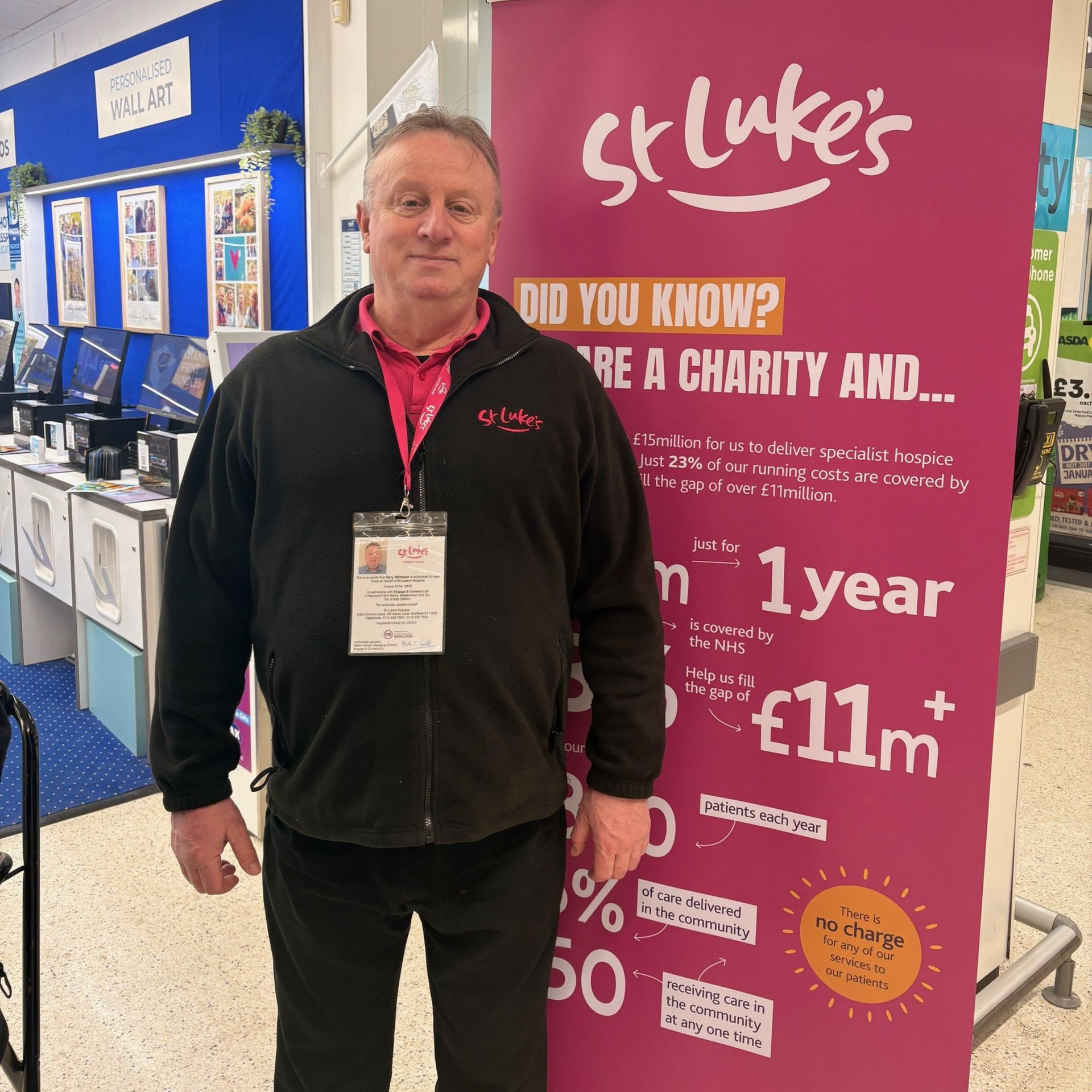 Man in a black fleece stands next to a St. Luke's charity banner in a store, displaying statistics.