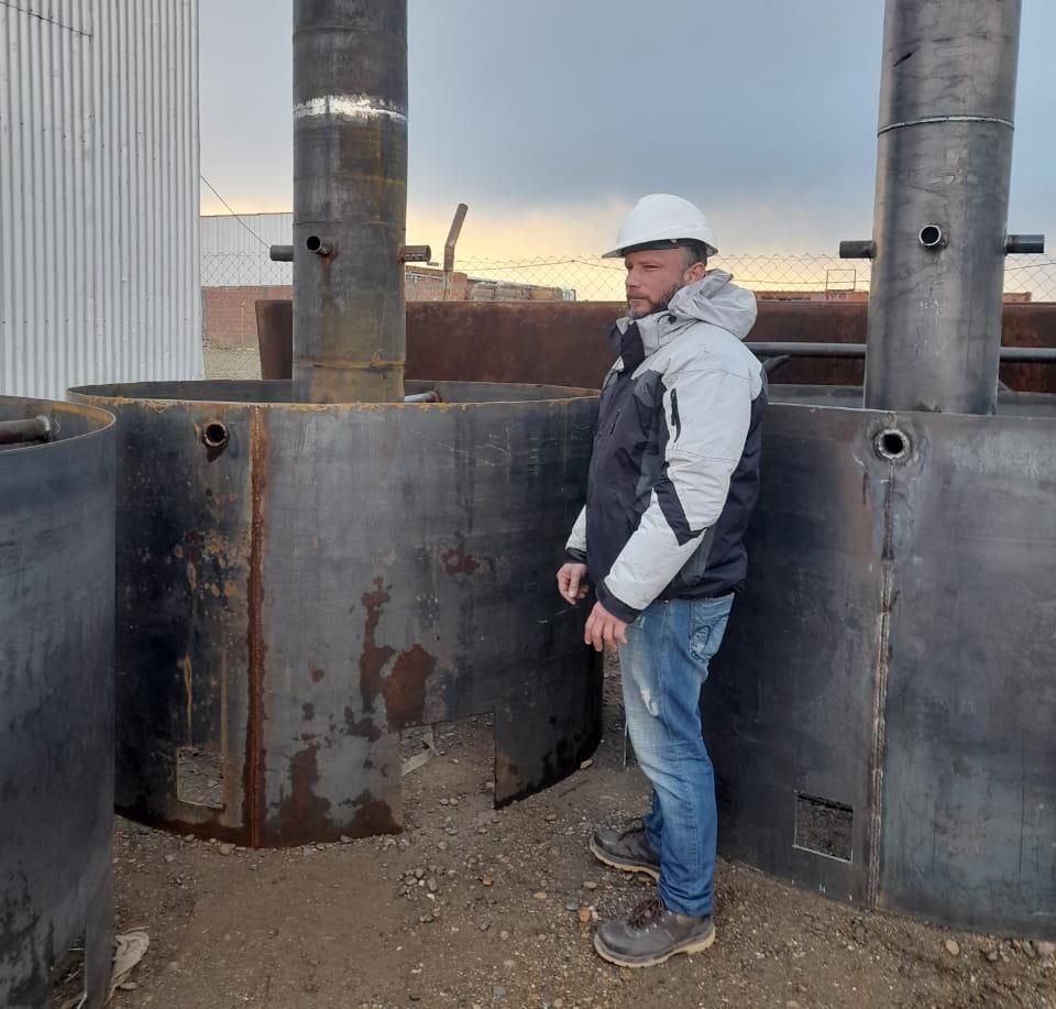 Un hombre con casco de seguridad se encuentra cerca de grandes estructuras metálicas cilíndricas al aire libre. Día nublado.