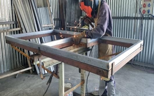 Un soldador con máscara y equipo de protección suelda un marco de metal en un taller.