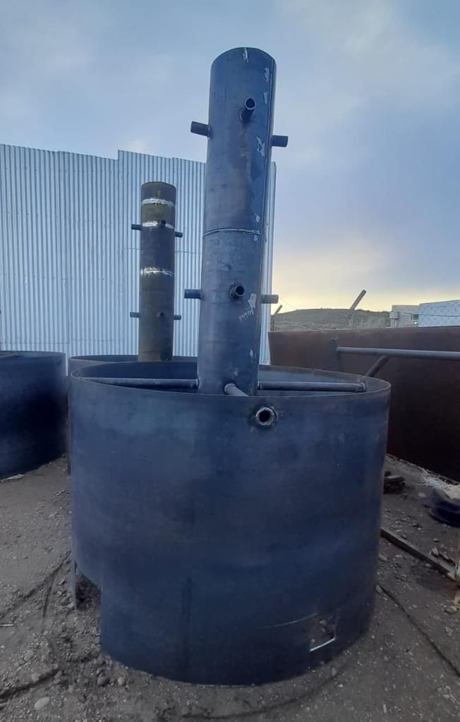 Tanque cilíndrico grande y oscuro de metal con un tubo vertical adjunto; al aire libre.