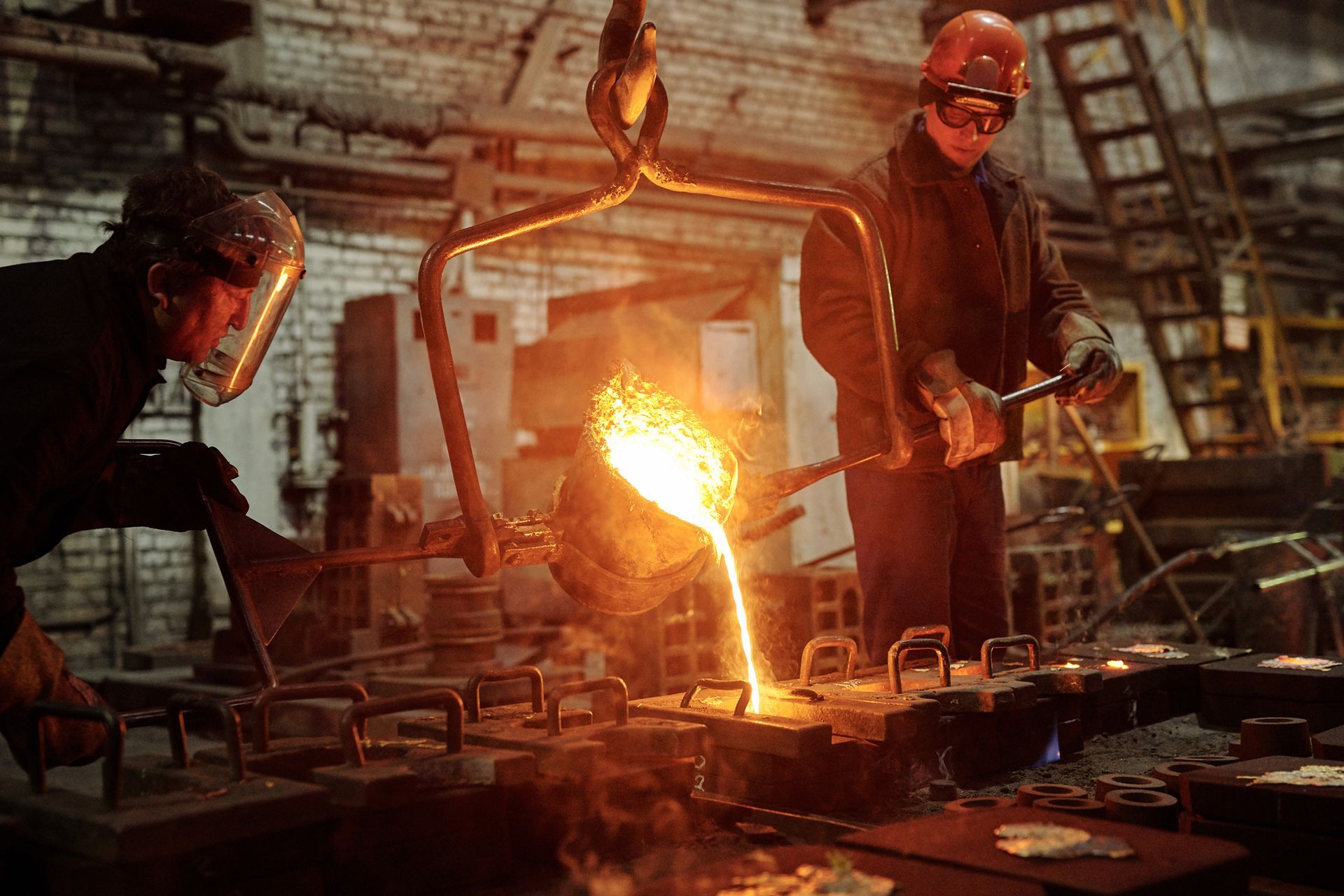 Trabajadores vierten metal fundido desde un crisol en moldes en una fundición, usando equipo de protección.