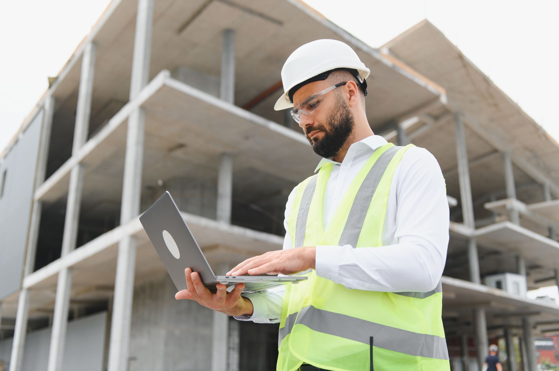 Construction worker in hard hat and safety vest using a laptop at a building site