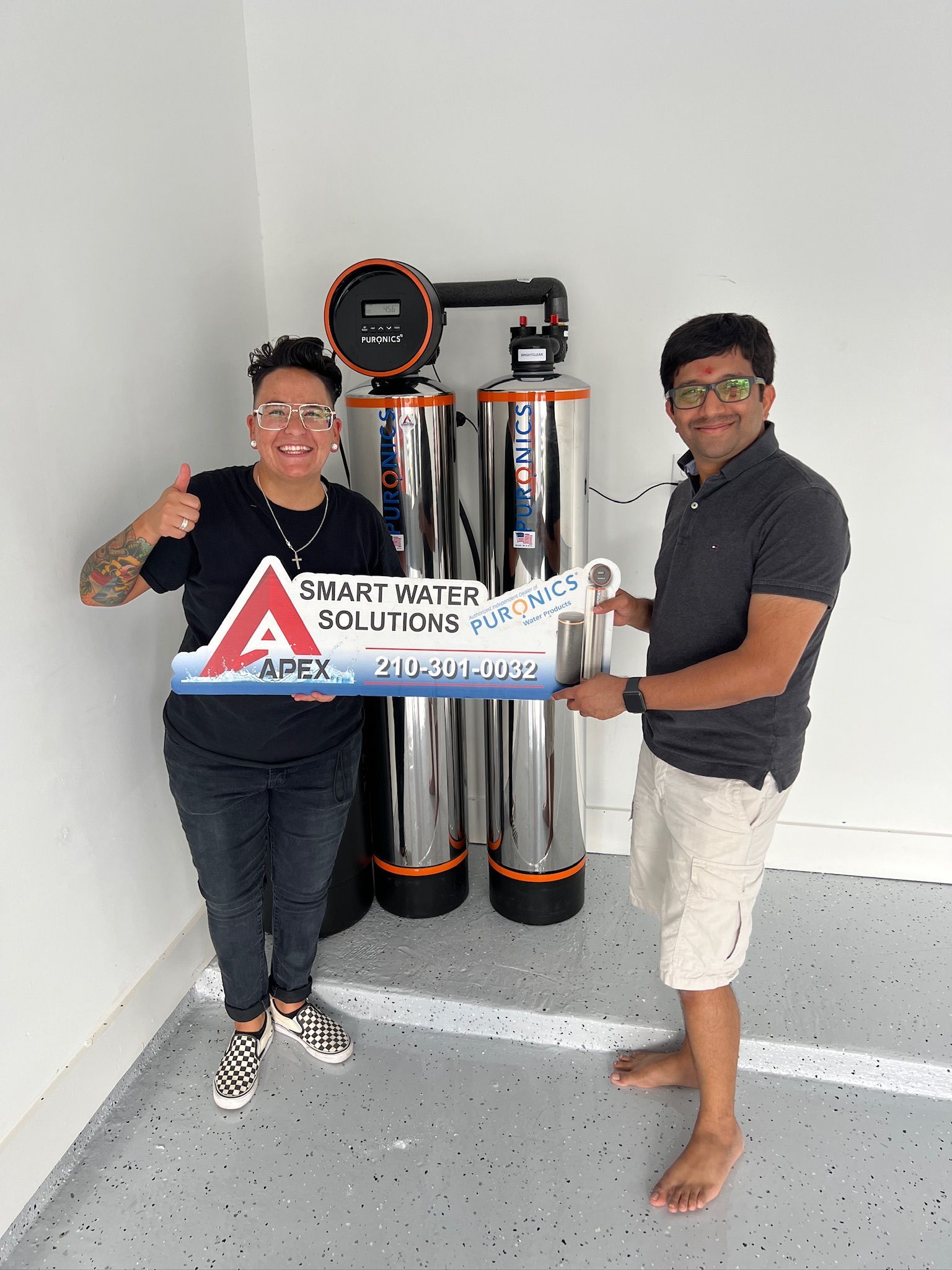 Two people holding a sign beside metal cylinders in a room, one giving a thumbs-up.