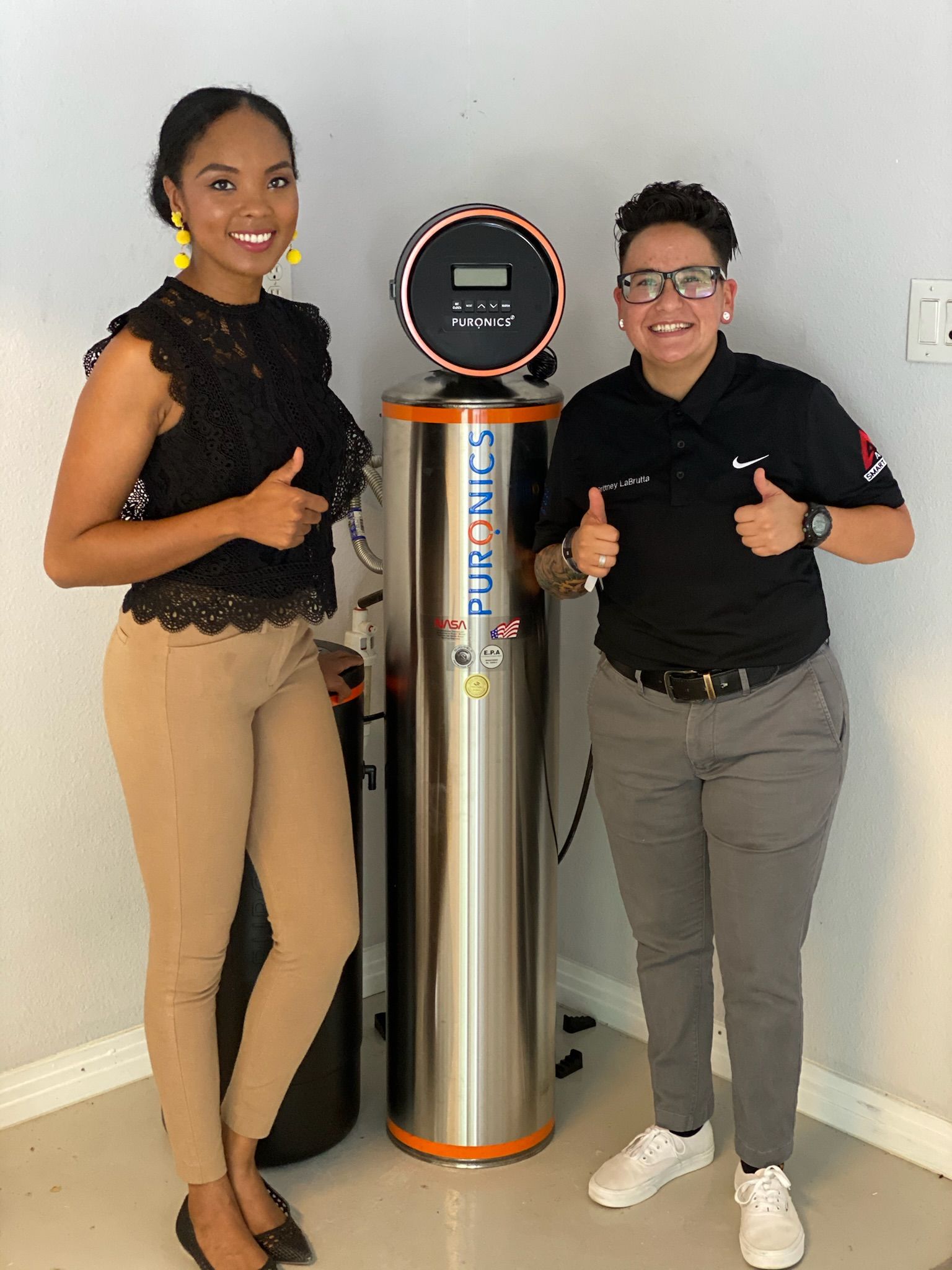 Two people posing with thumbs up beside a tall black-and-silver device in a room