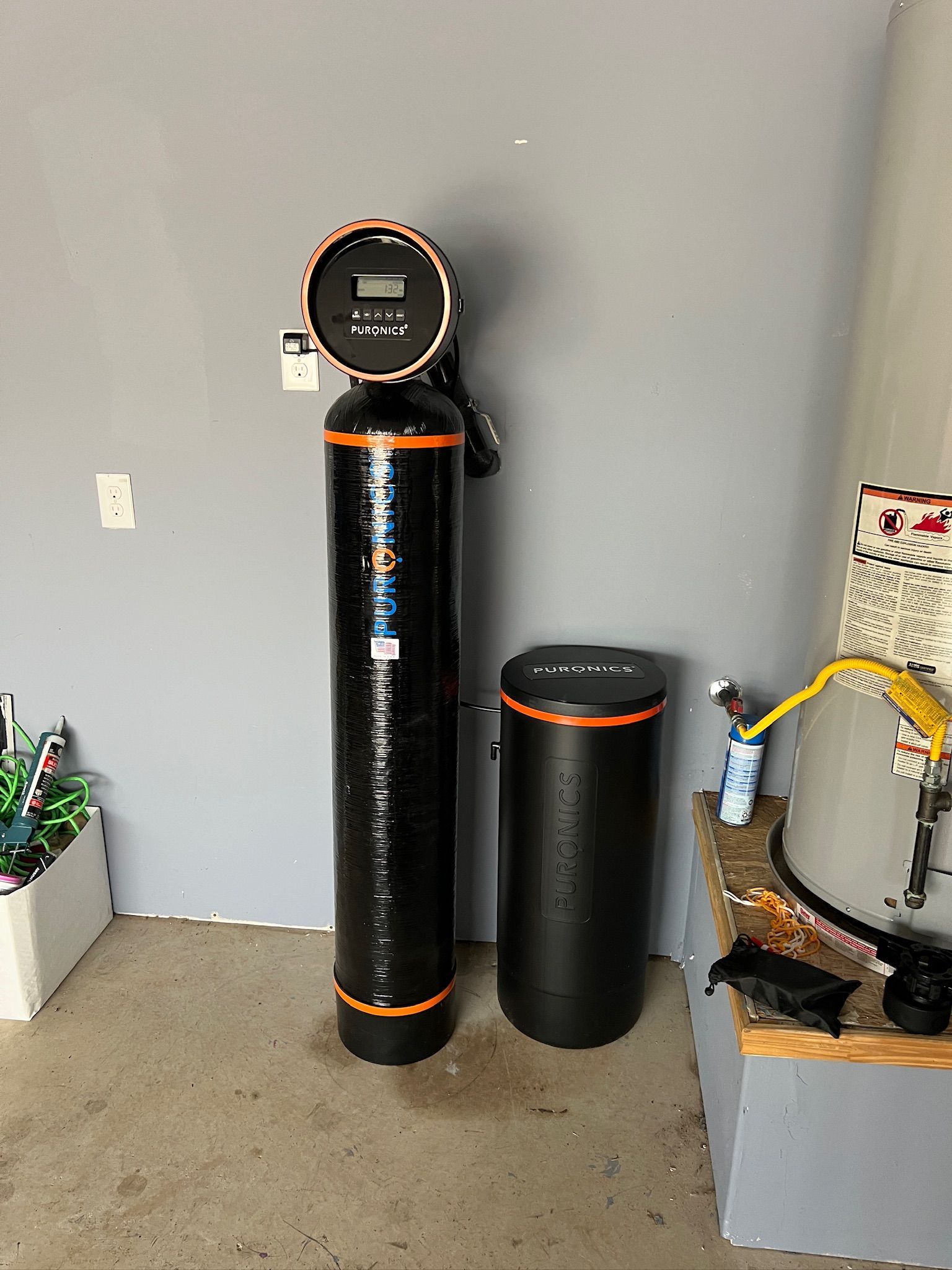 Black water softener tank and smaller cylinder in a utility room corner next to piping and a bucket