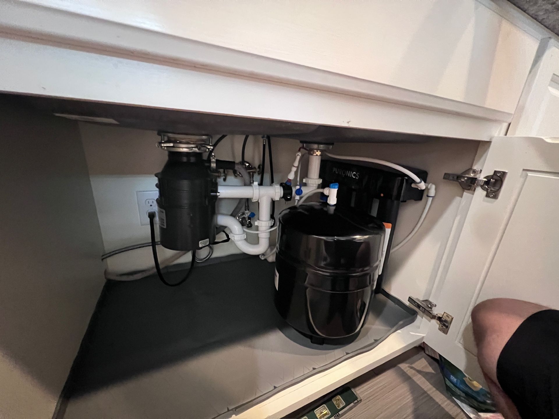 Under-sink cabinet with exposed plumbing, a black garbage disposal, and a large black trash can.