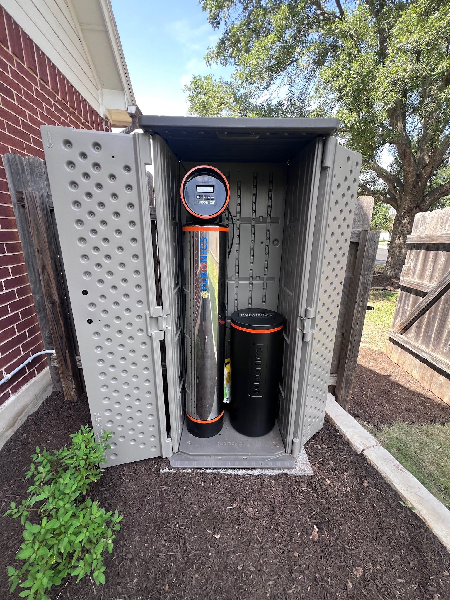 Outdoor utility enclosure with gas meter and black tank beside a brick house and fence.