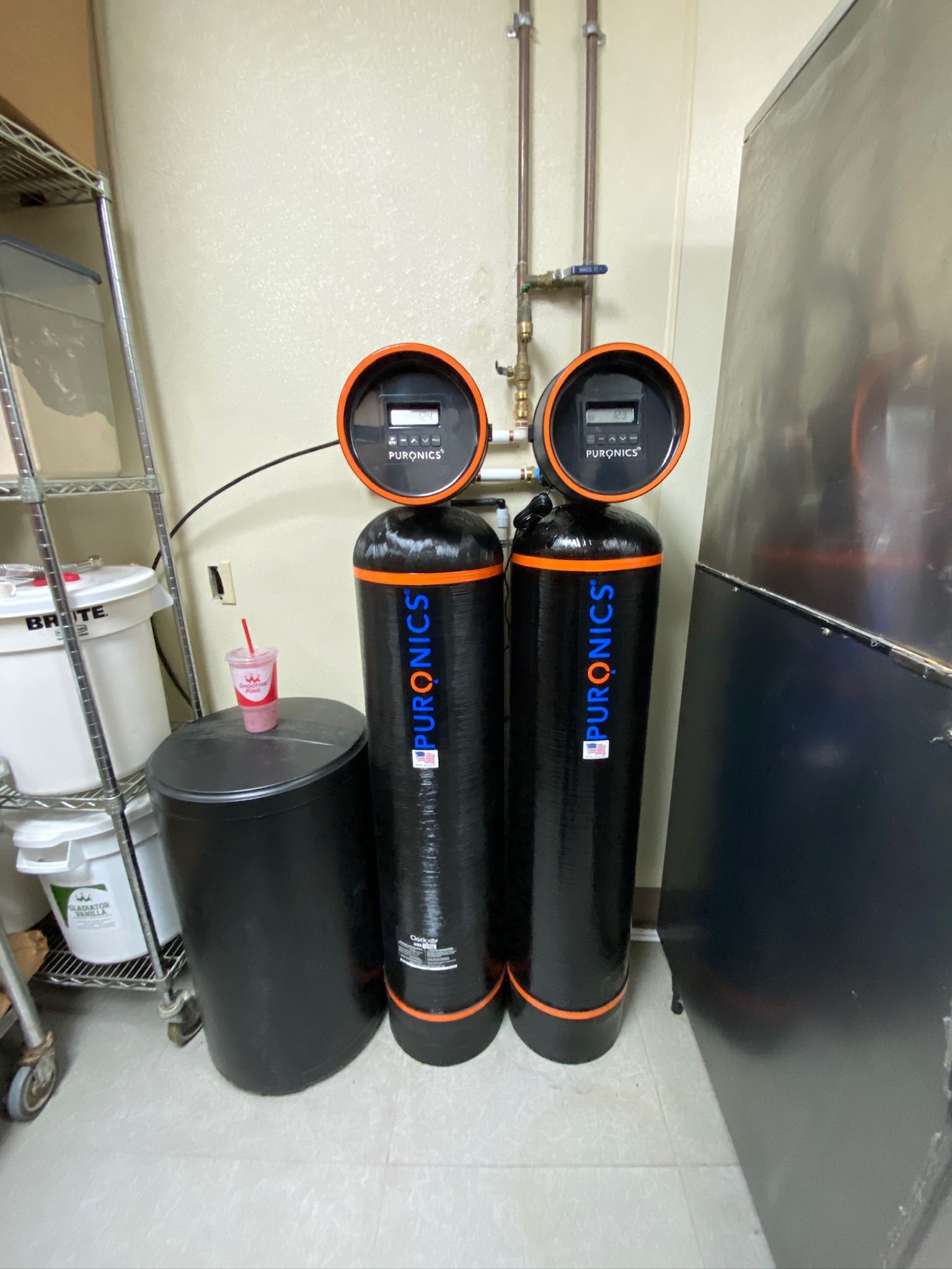 Two black water softener tanks with control heads beside a gray utility tank in a basement utility room