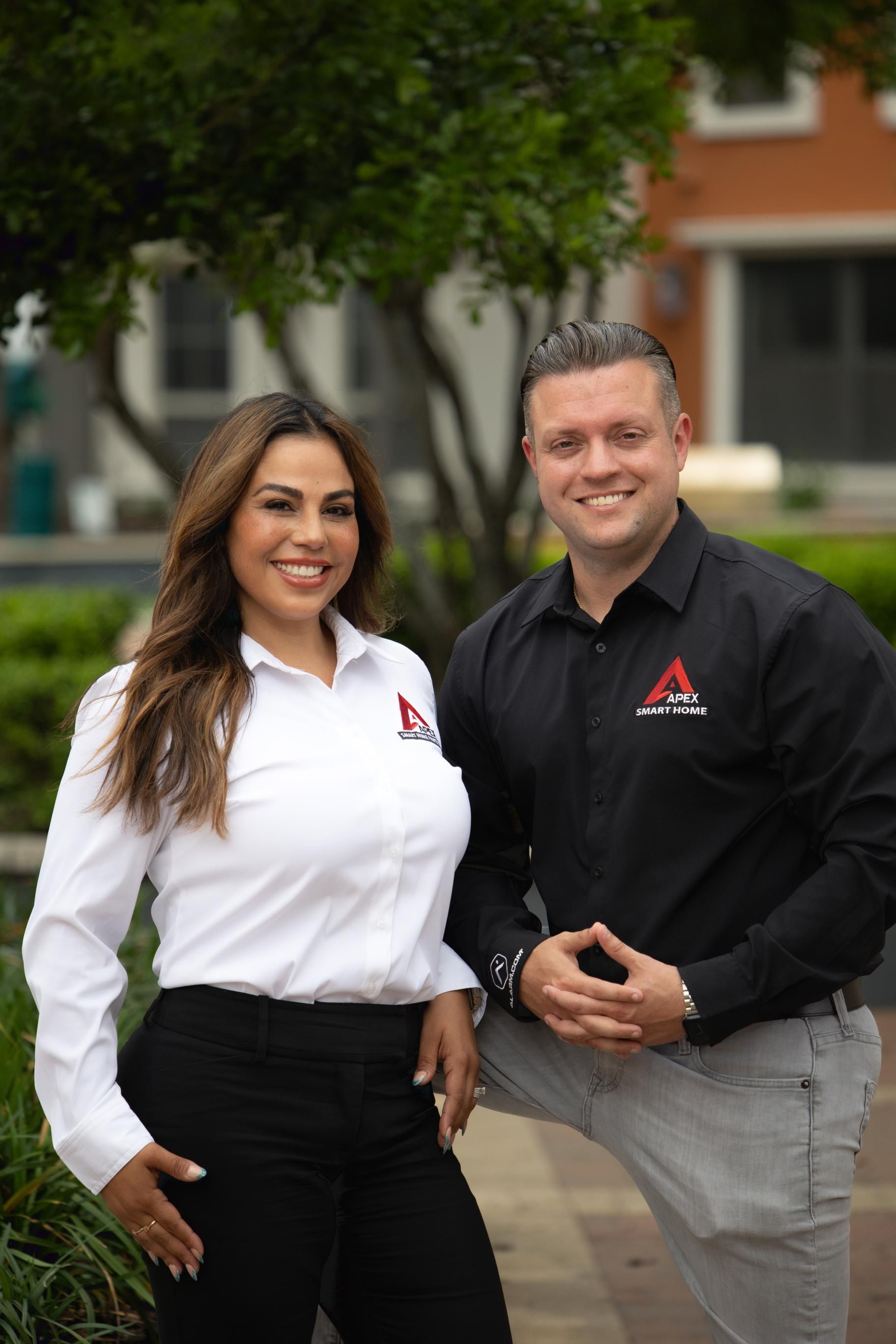Two people in branded shirts standing outdoors, smiling on a walkway in front of a building.