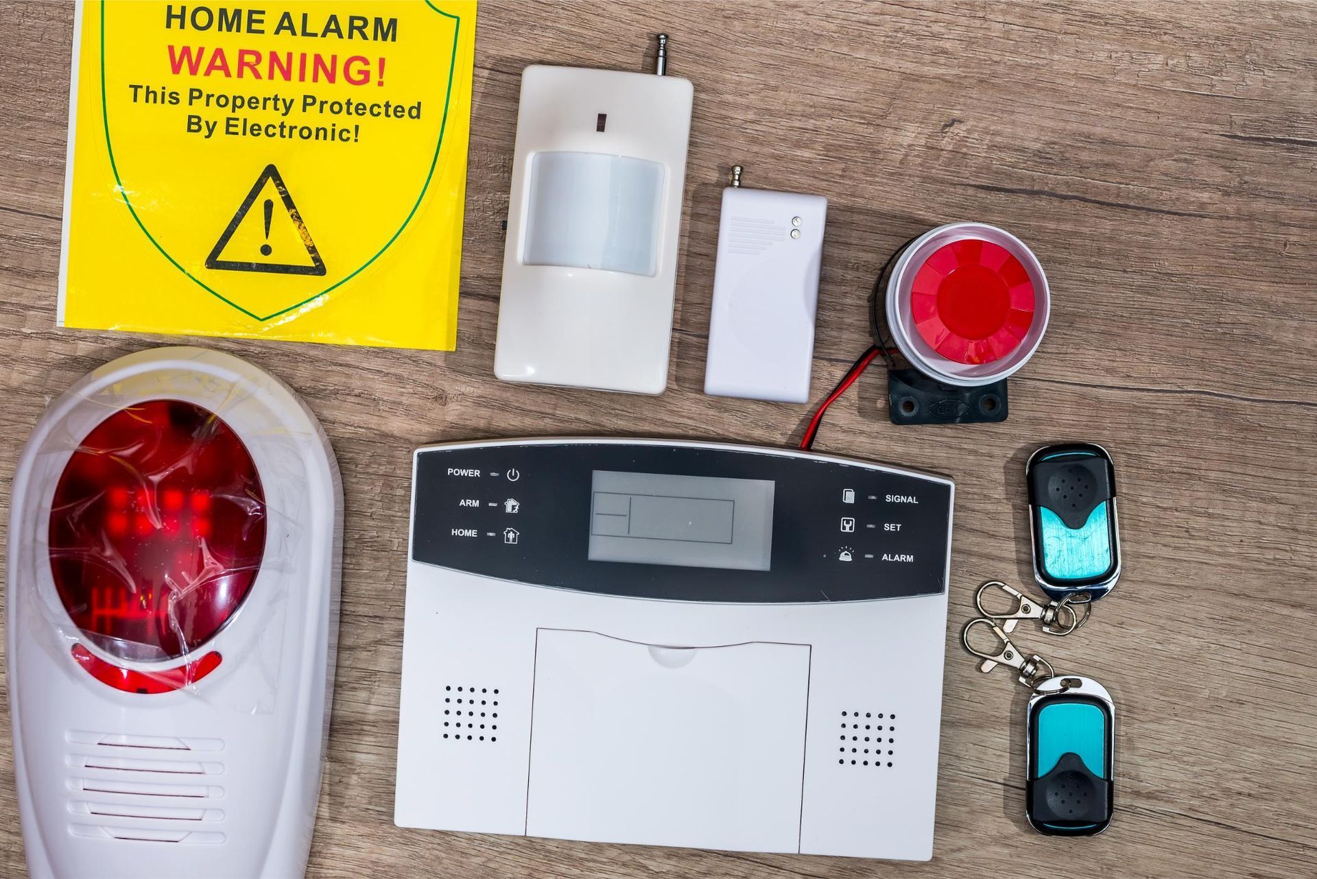 Home alarm components on a table: siren, keypad panel, sensors, remote fobs, and warning sign.