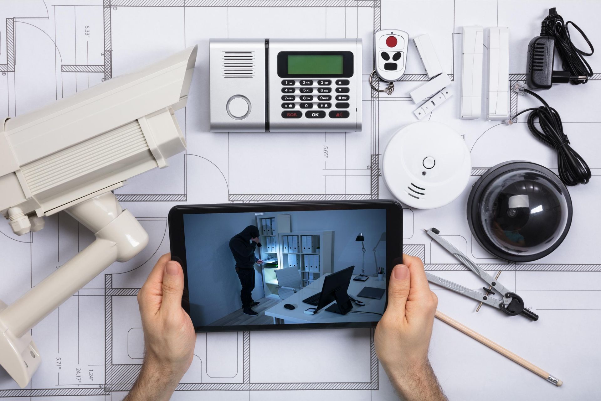 Hands holding a tablet showing a home security camera feed beside surveillance devices on a tiled floor