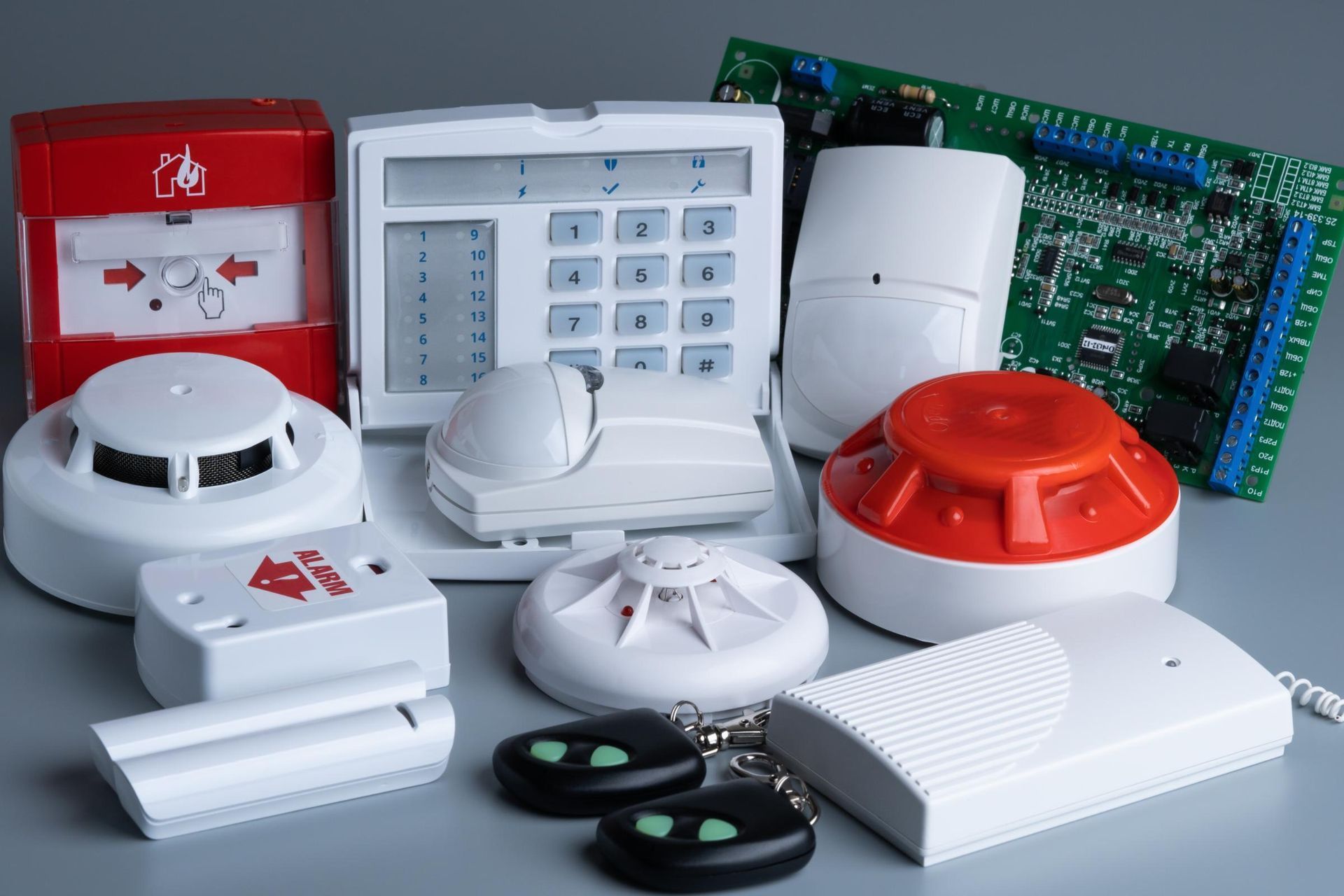 Assorted home security alarm devices and sensors in white, red, and green on a gray surface