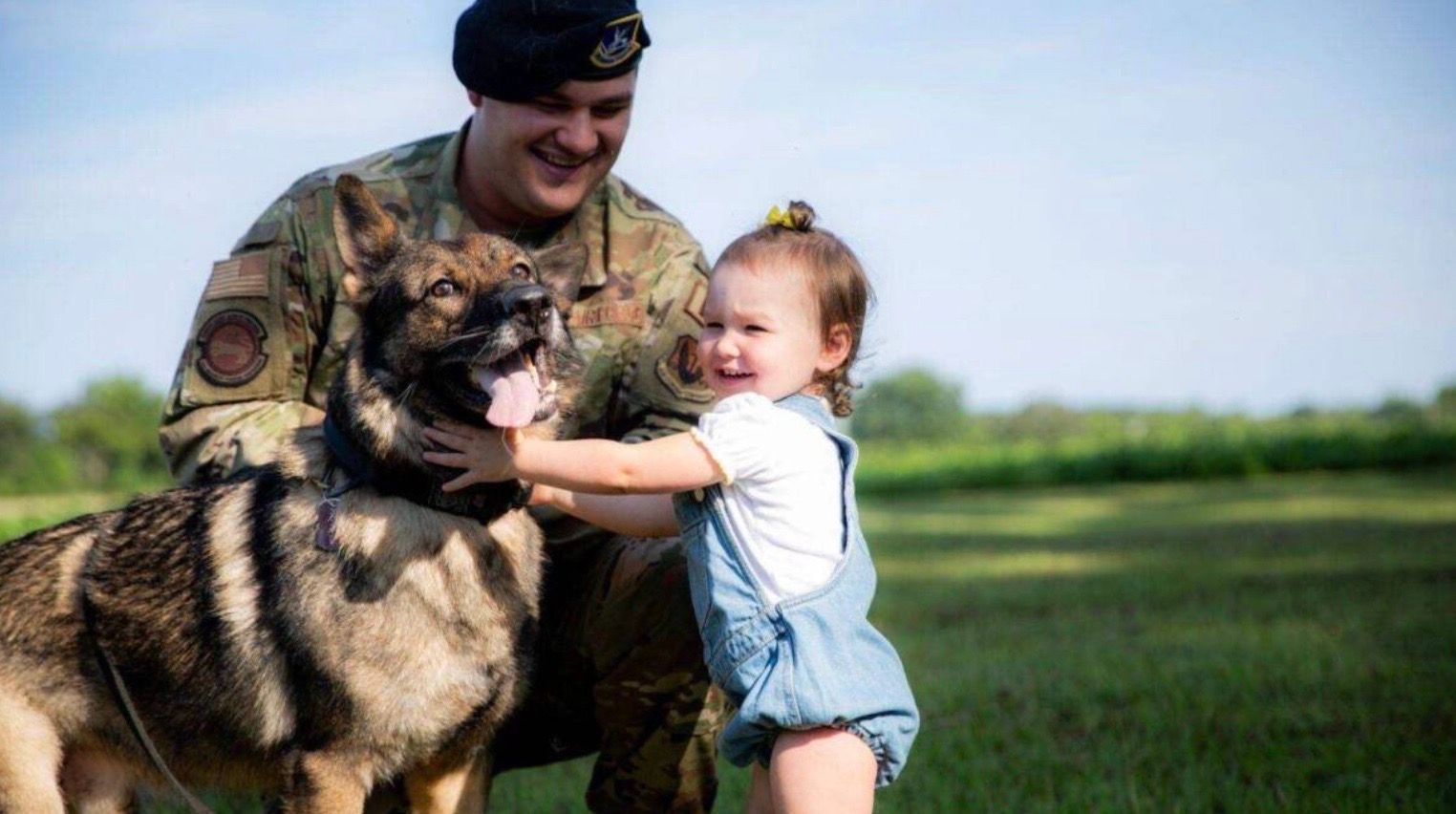 A soldier and a little girl are petting a dog in a field.