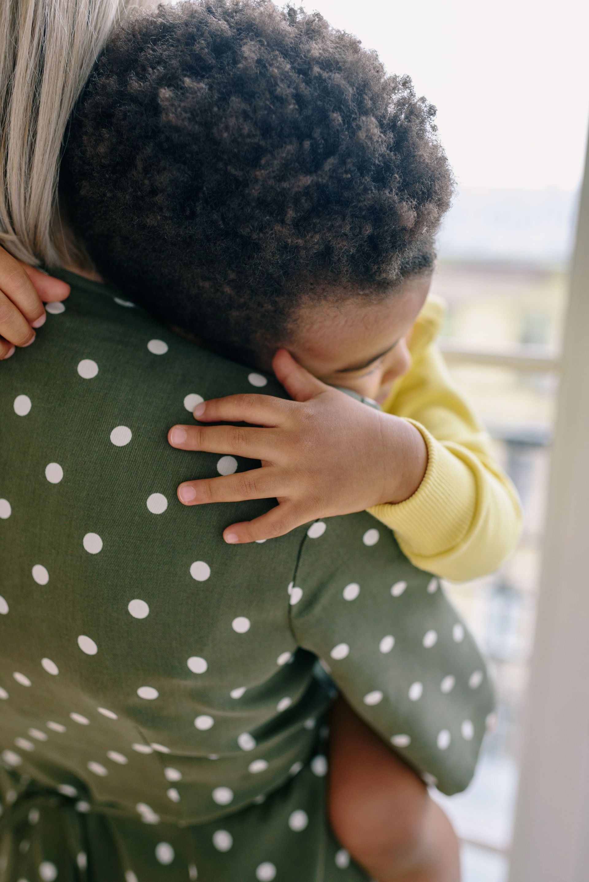 Child hugging an adult wearing a green polka-dot shirt indoors by a window