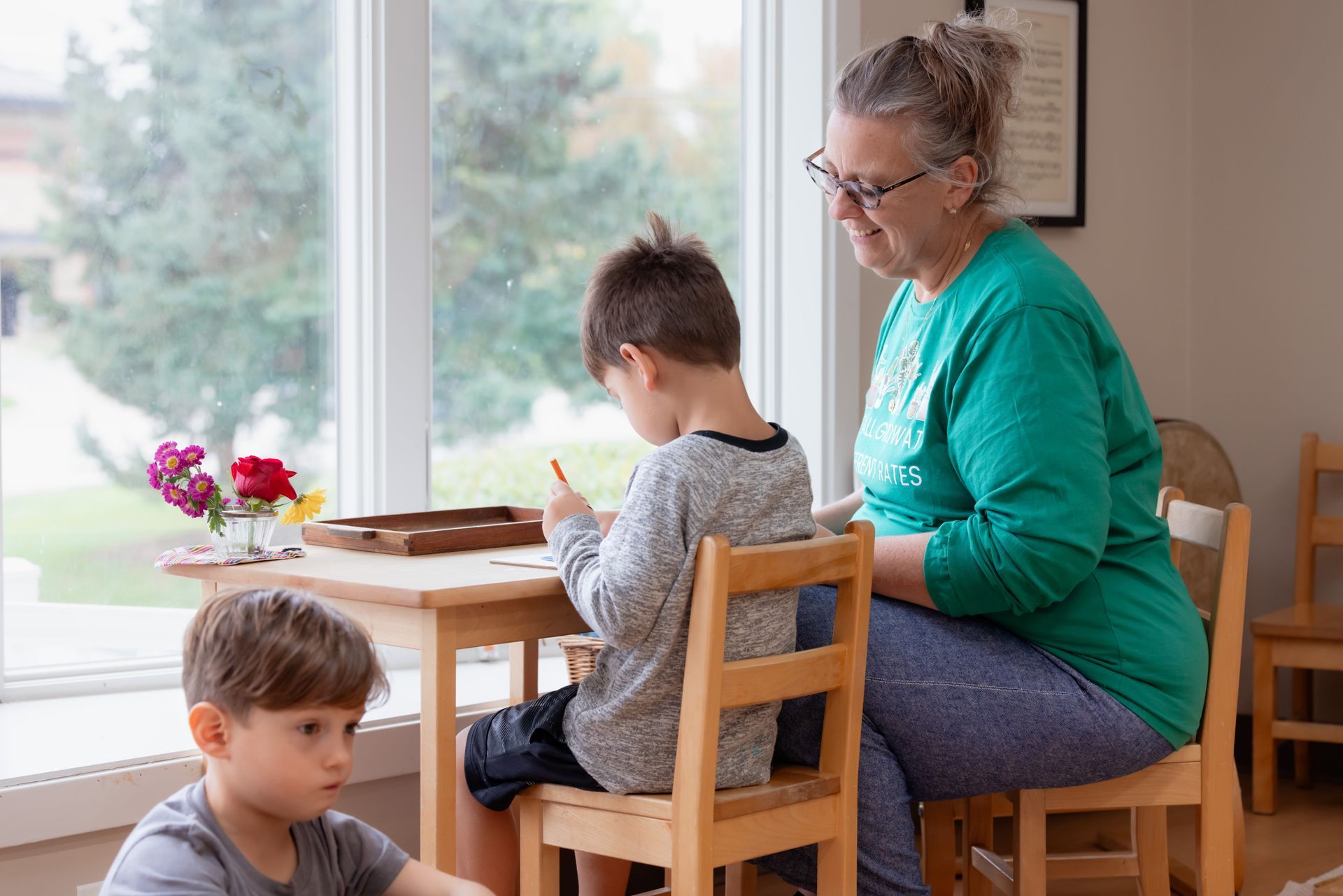 A child and a teacher sit at a wooden table by a bright window, doing work together