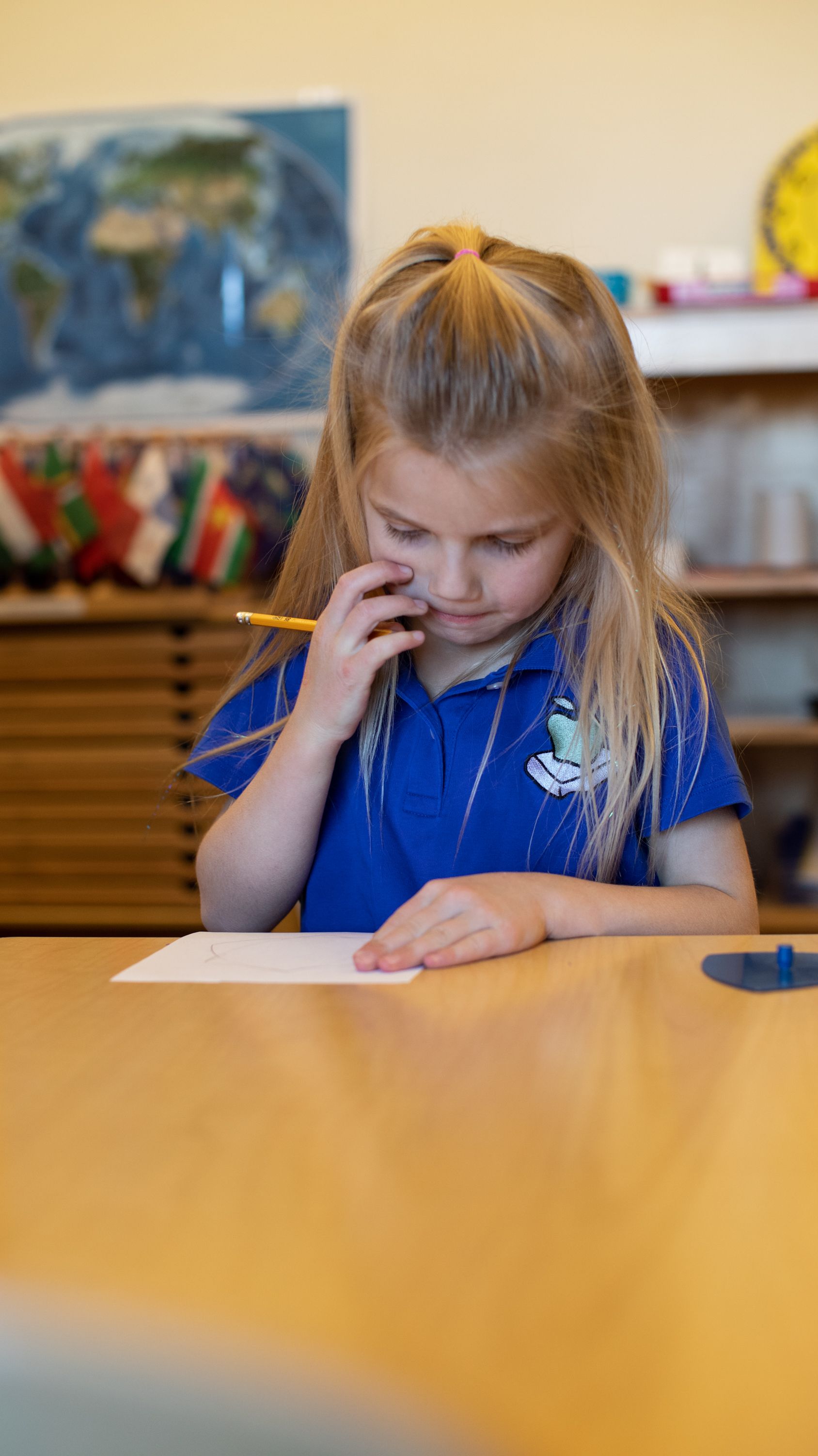 Young child in a blue sweater writing at a table in a classroom, focused on a metal inset