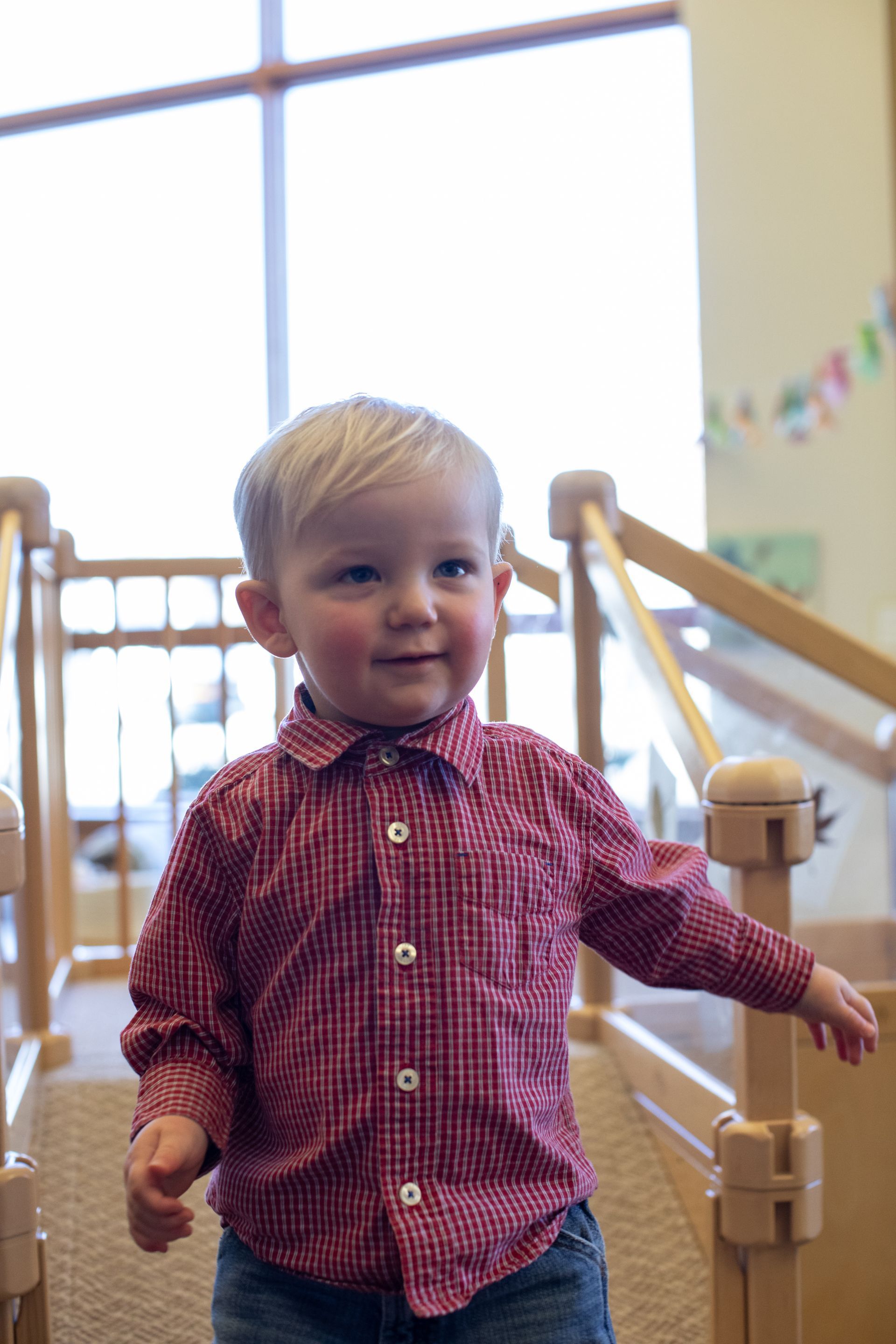 Toddler in a red checkered shirt walking indoors beside a wooden railing