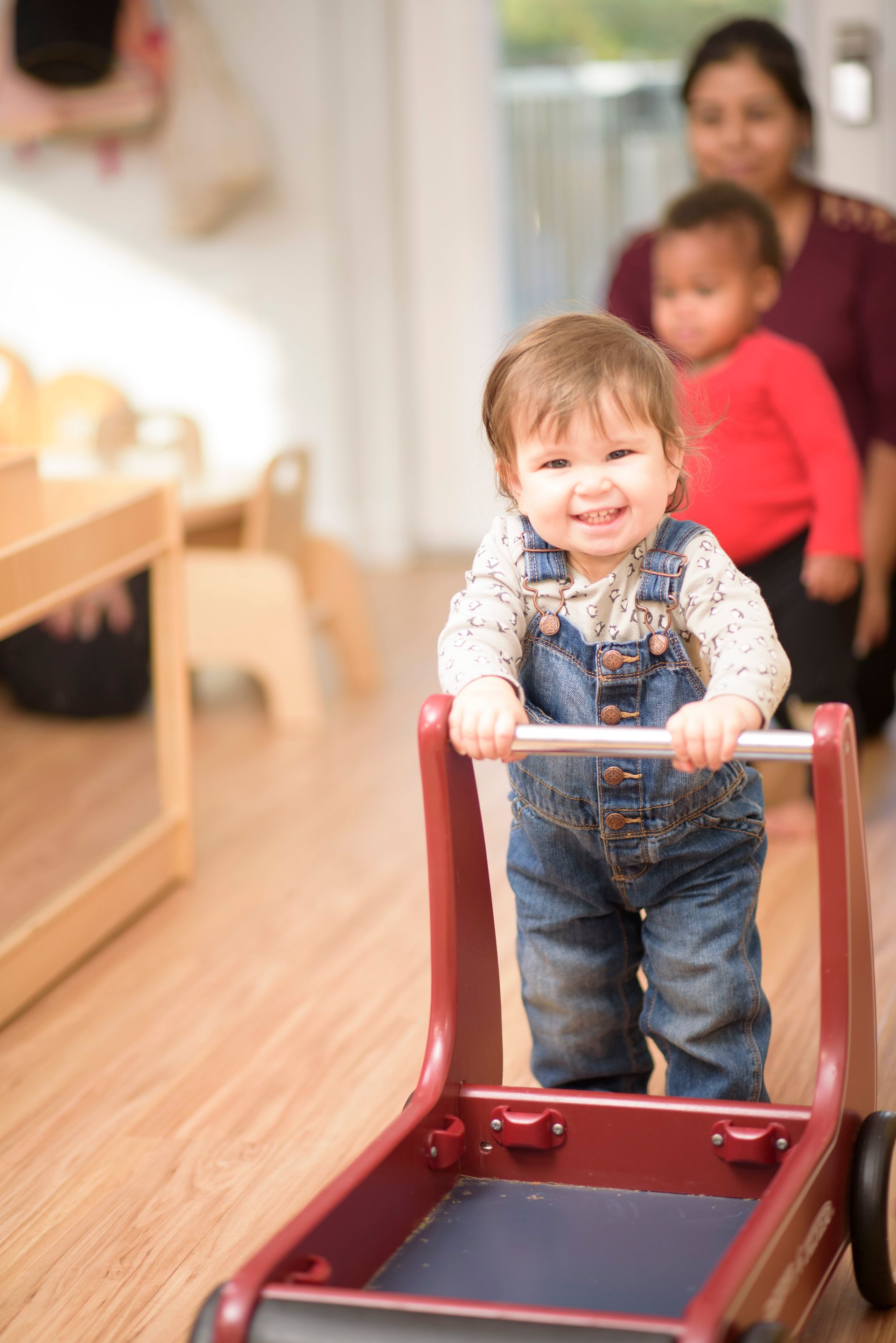 Toddler smiling while pushing a red toy cart indoors, with an adult and child in the background
