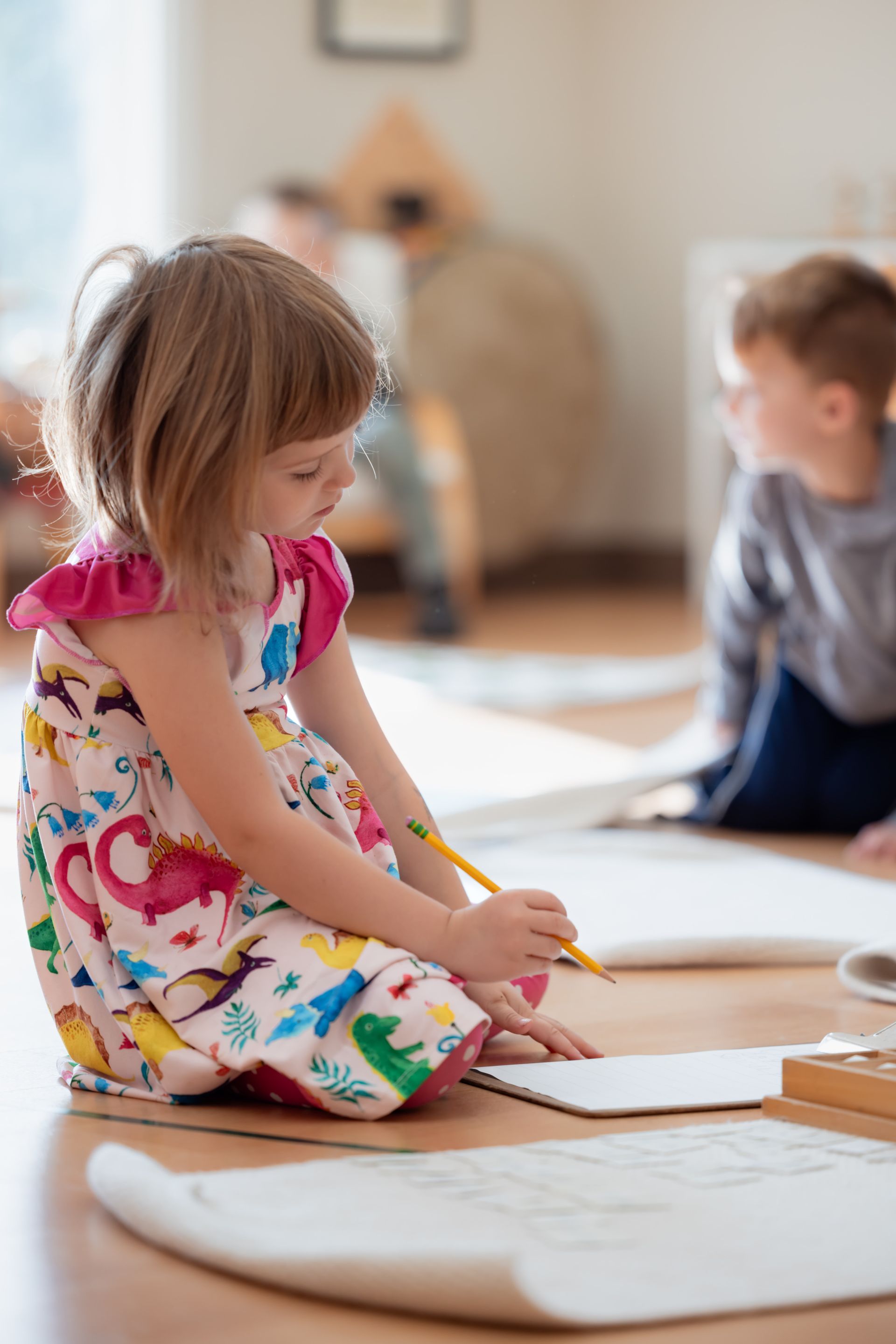 Child writing on paper in a bright classroom, with another child blurred in the background