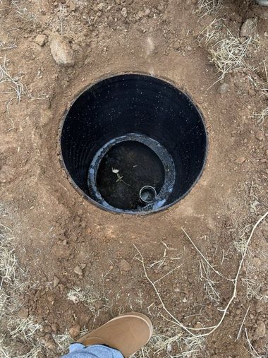 Open black cylindrical tank set in dirt, viewed from above with a shoe at the bottom edge.