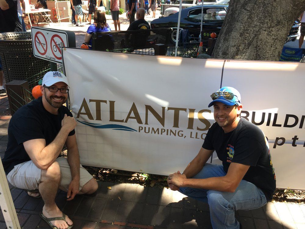 Two men crouch beside an Atlantis Plumbing sign outdoors under a tree.