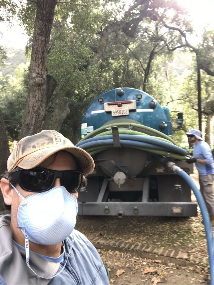 Person in cap and mask taking a selfie beside a blue tanker truck in a wooded area