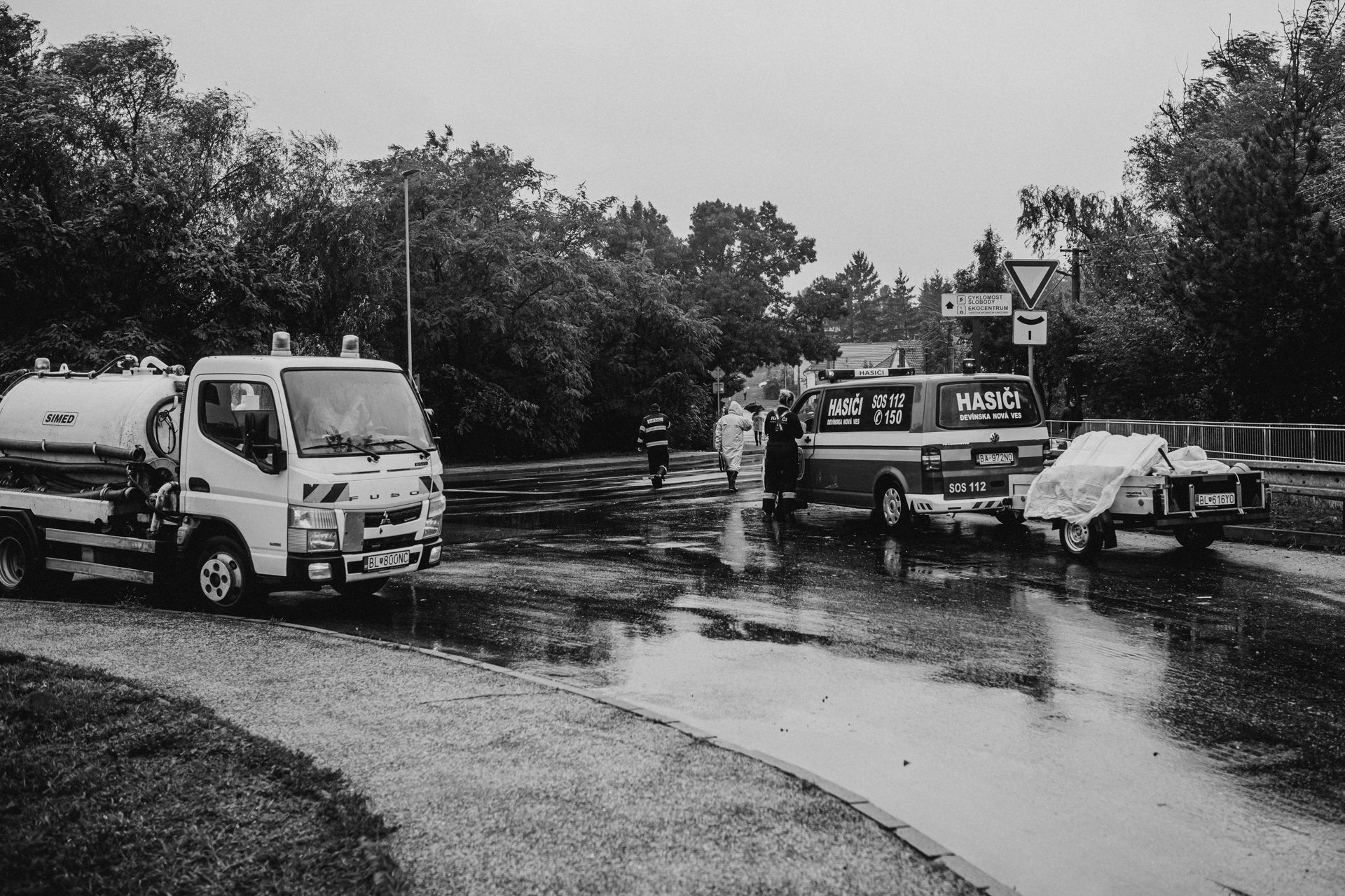 Rainy roadside scene with a tanker truck, a van, and people beside parked vehicles