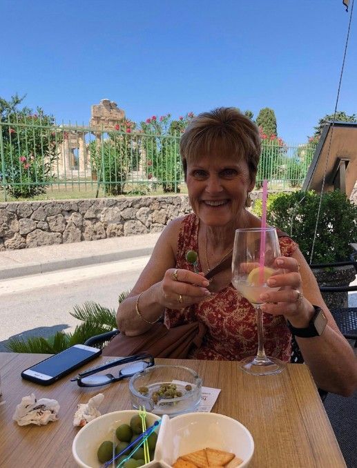 Woman smiles, holding a cigar and drink, seated at outdoor cafe with stone building backdrop.