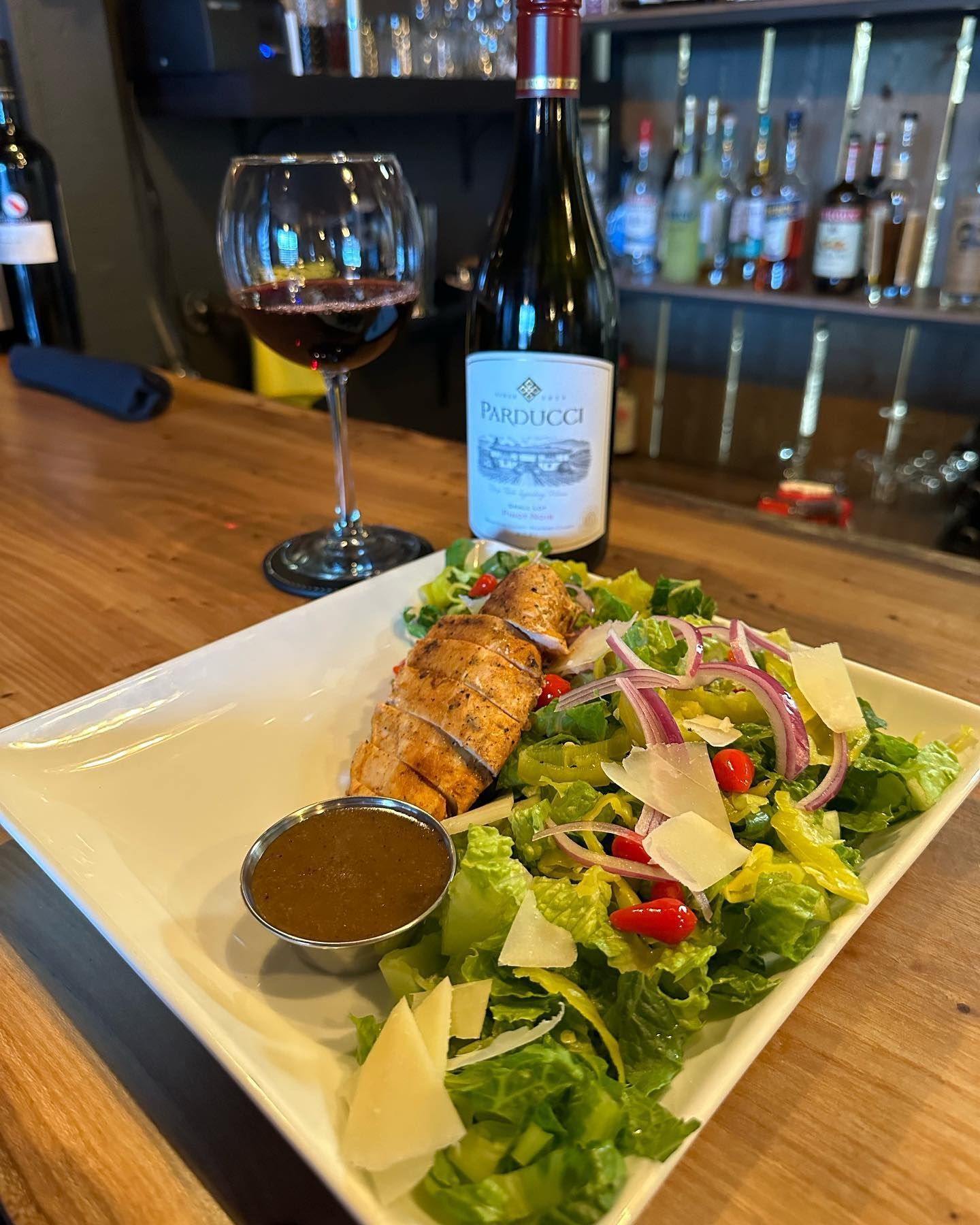 Grilled chicken Caesar salad, wine glass, and bottle of wine on a wooden bar.