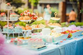 Outdoor party table with desserts, sandwiches, and flowers; people in background.