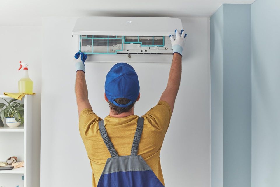 A technician in a blue cap and work overalls cleaning the air filters of a white wall-mounted air conditioner.