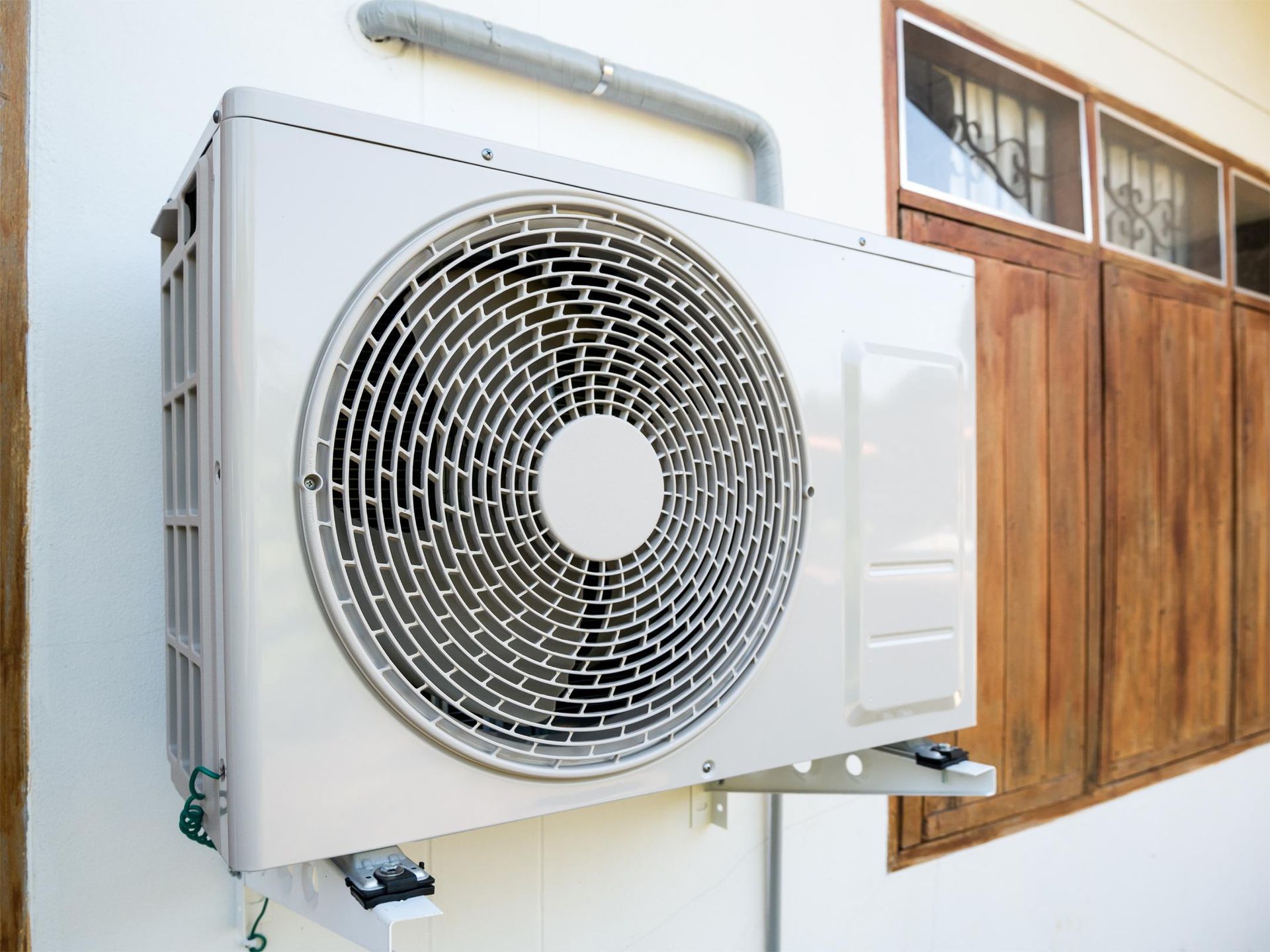 A white air conditioning condenser unit mounted on an exterior wall next to a wooden-framed window.