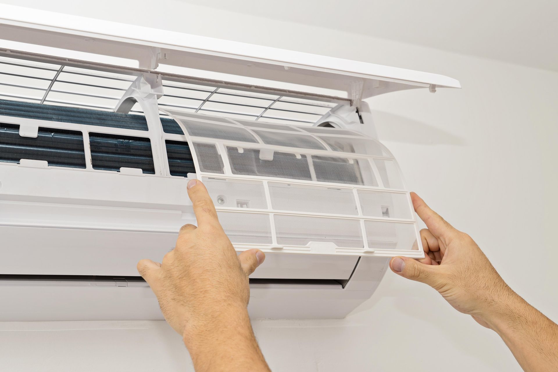 Hands removing a clean white mesh filter from an open wall-mounted air conditioning unit.