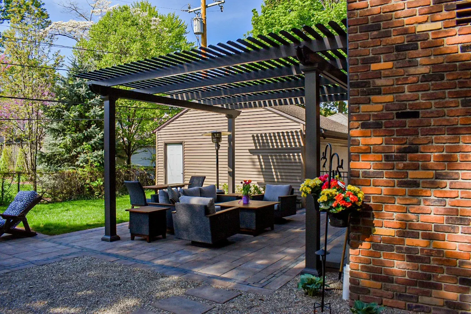 A circular stone fire pit sits on a flagstone patio in a backyard with a curved stone retaining wall and green lawn.
