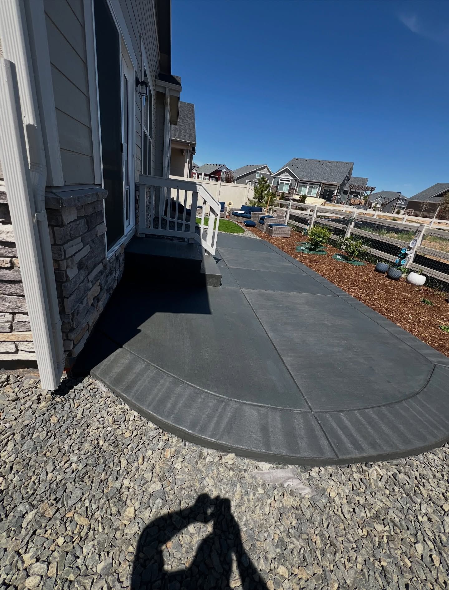 A newly installed, dark gray concrete patio with a rounded edge next to a home with stone siding and a white staircase.