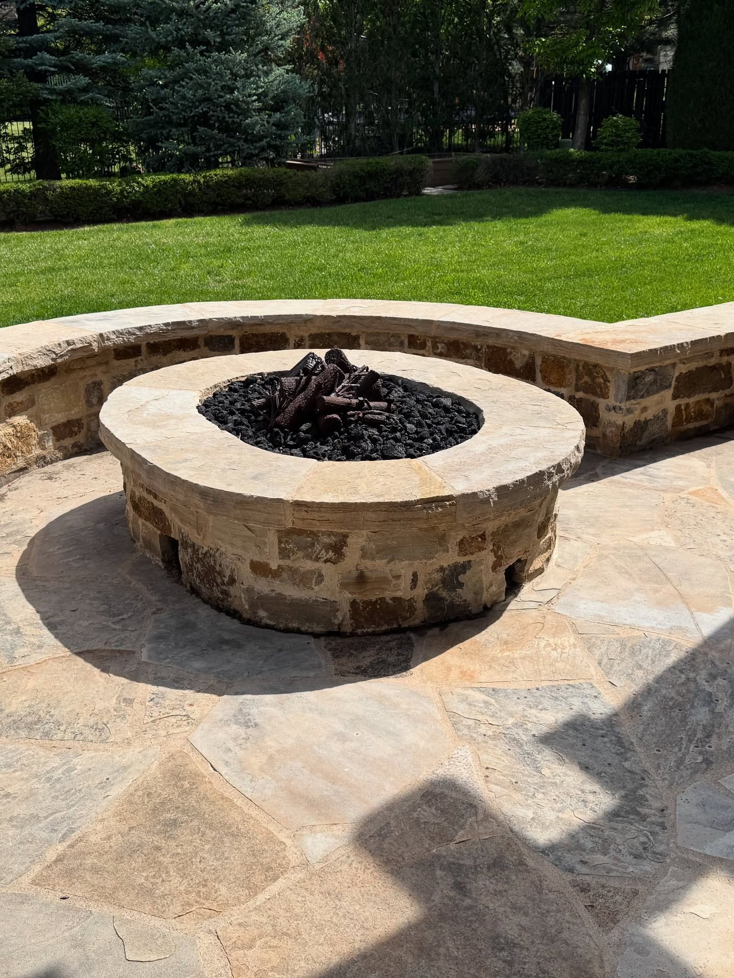 A circular stone fire pit sits on a stone patio in front of a curved stone retaining wall and a green lawn.