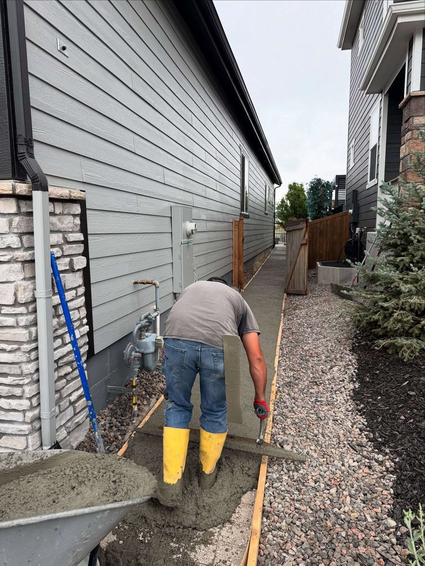 A person in yellow boots smooths wet concrete into a narrow walkway between two houses.