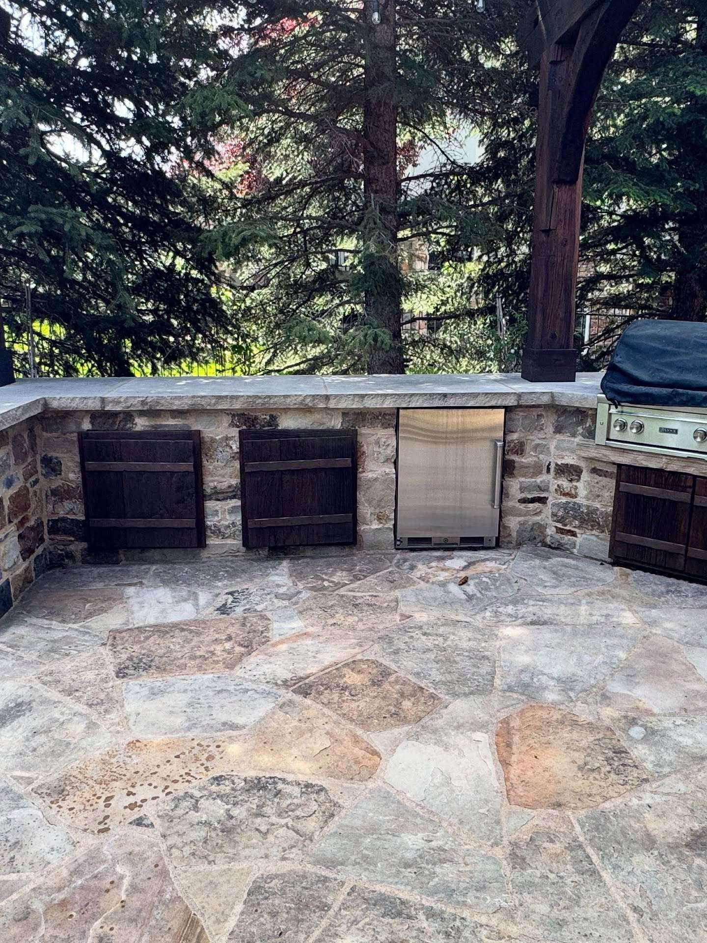 An outdoor stone kitchen area with a built-in stainless steel refrigerator and dark wooden cabinets under a stone counter.