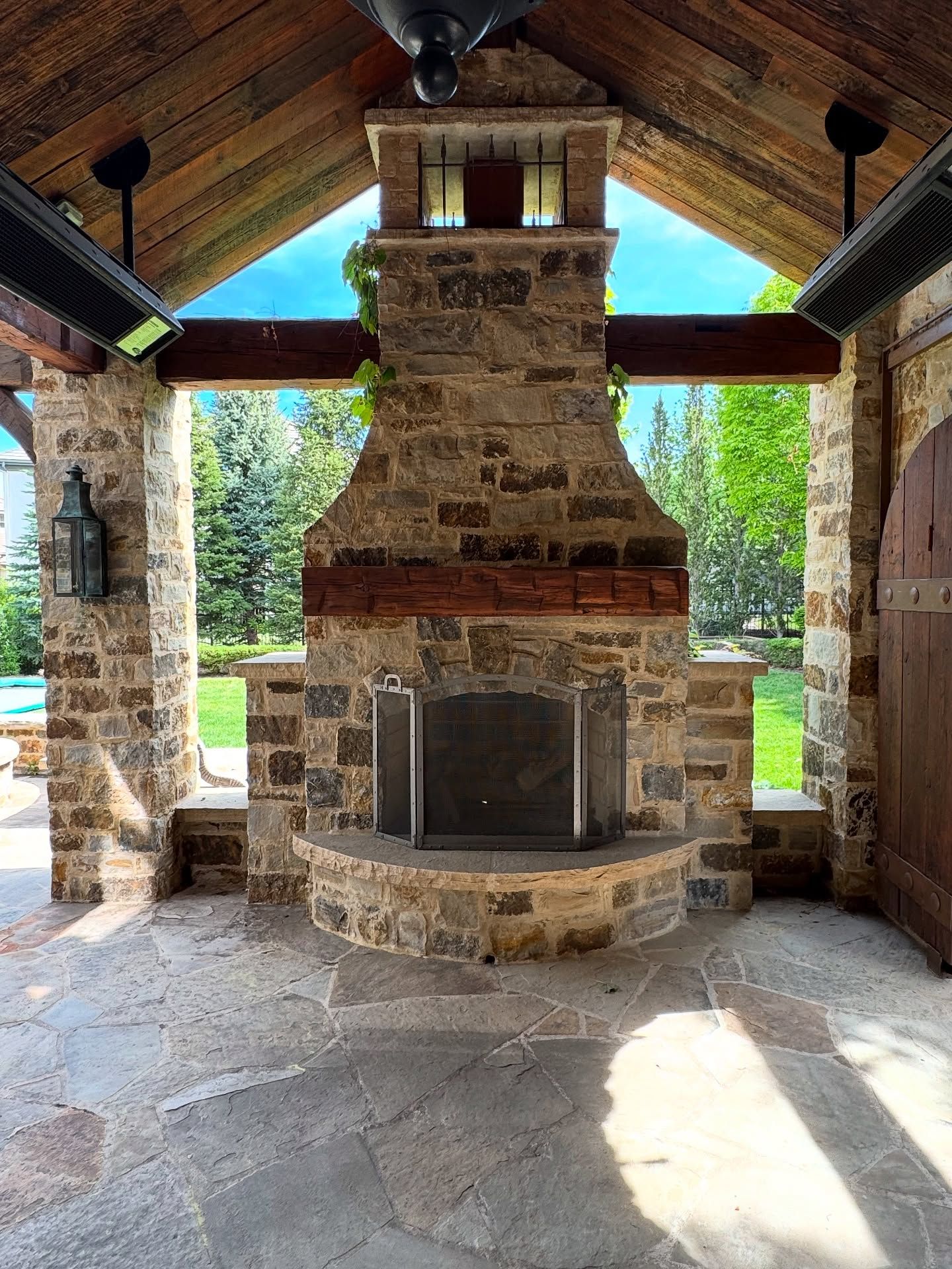 A stone outdoor fireplace under a rustic, open-air wooden patio structure with a flagstone floor and garden backdrop.