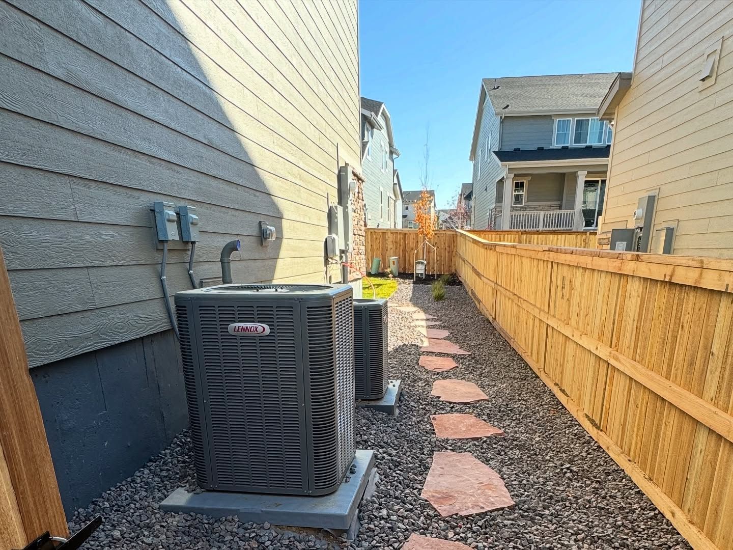 Side yard of a house featuring a grey AC unit on gravel, a stone path, and a wooden privacy fence under a clear blue sky.