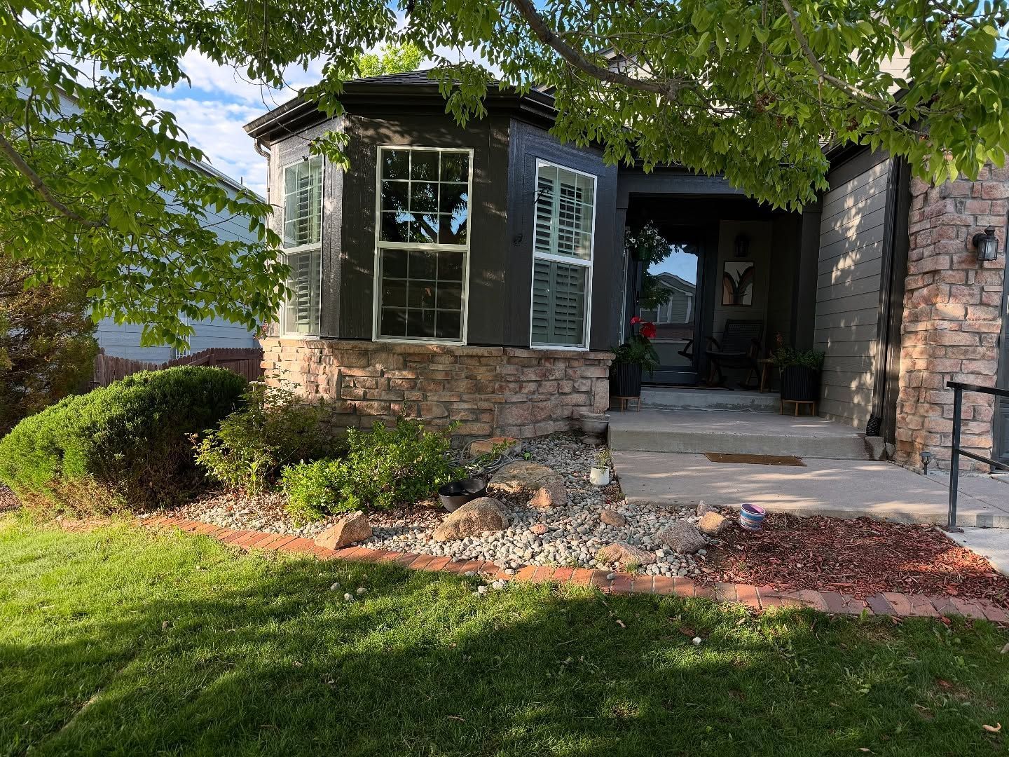 A residential home exterior featuring a bay window, stone foundation, and landscaped yard with green grass and rock beds.