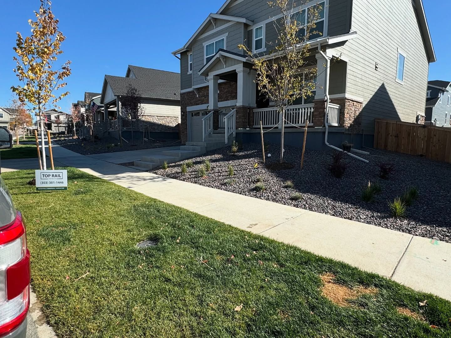 A suburban street with multi-story houses, manicured lawns, young trees, and a sidewalk under a clear blue sky.