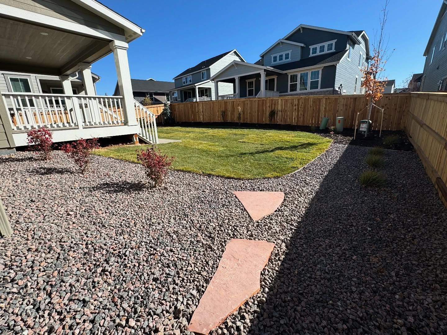 A backyard with a gravel area, stone path, a small patch of grass, and a wooden fence, seen from a patio deck.